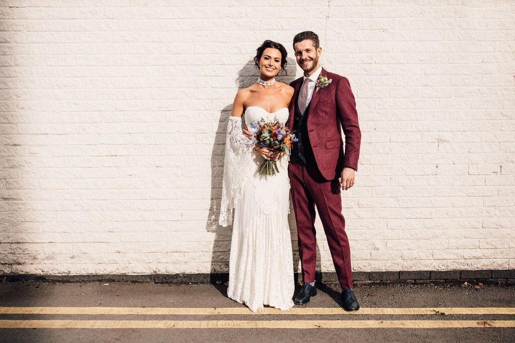 A happy bride and groom pose in front of a brick wall, with the groom wearing a maroon suit.