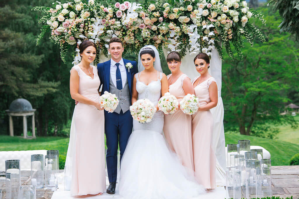 The bride and groom pose with the bridesmaids in front of their flower arch.