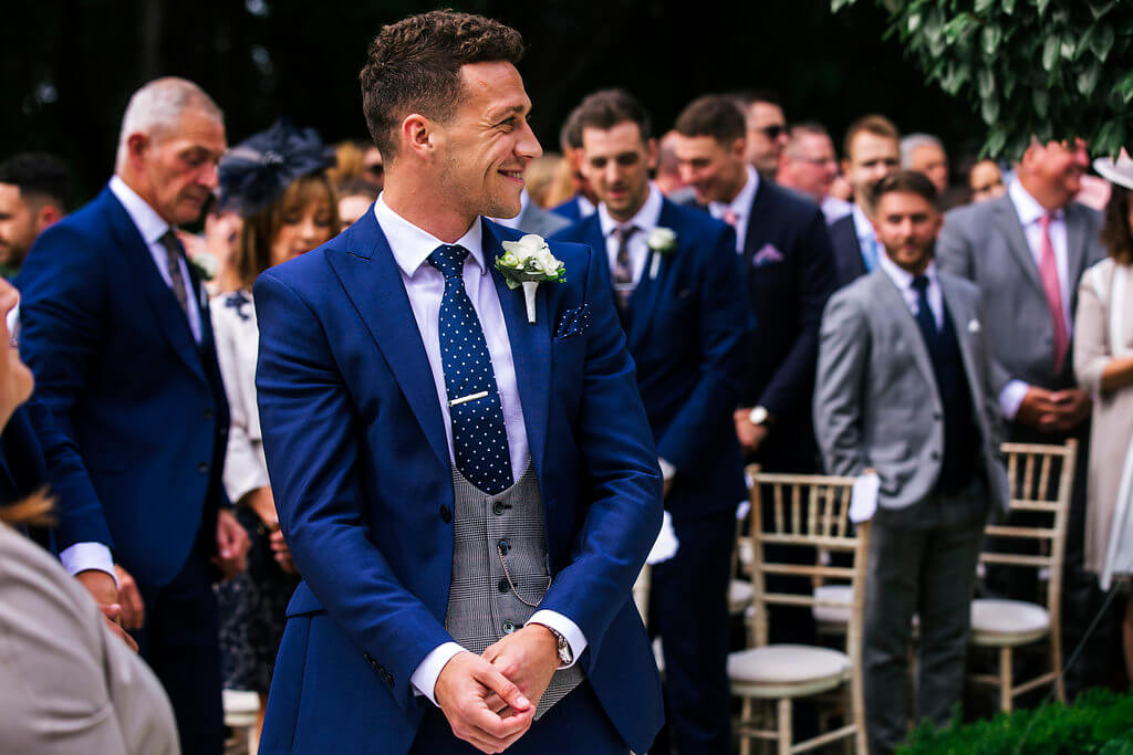 A candid shot of the groom smiling as he waits for his bride. The groom is wearing a 3 piece navy suit with a grey checked waistcoat.