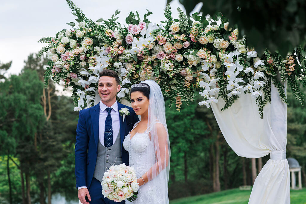 The bride and groom standing by an arch of flowers. The groom is wearing a navy suit with a grey waistcoat, with the bride holding her bridal bouquet.