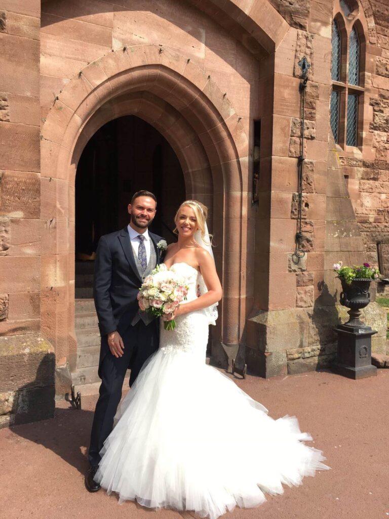 Bride and groom smiling at the camera stood outside of Peckforton Castle. Bride is holding a bridal bouquet.