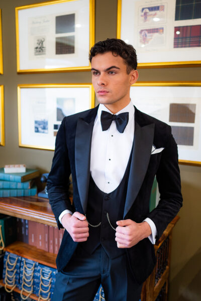 Slim fit classic black velvet dinner suit with traditional black velvet tux jacket, classic black dinner suit trousers and black u shape horseshoe waistcoat. Features a slim fit 1 button velvet dinner jacket with wide black satin peak lapel. Worn with white studded dress shirt with French cuff shirt and large tom ford style bow tie and white pocket square.