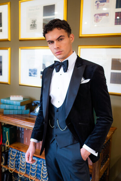 Slim fit classic black velvet dinner suit with traditional black velvet tux jacket, classic black dinner suit trousers and black u shape horseshoe waistcoat. Features a slim fit 1 button velvet dinner jacket with wide black satin peak lapel. Worn with white studded dress shirt with French cuff shirt and large tom ford style bow tie and white pocket square.