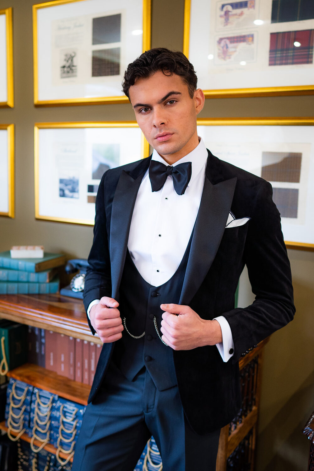 Slim fit classic black velvet dinner suit with traditional black velvet tux jacket, classic black dinner suit trousers and black u shape horseshoe waistcoat. Features a slim fit 1 button velvet dinner jacket with wide black satin peak lapel. Worn with white studded dress shirt with French cuff shirt and large tom ford style bow tie and white pocket square.
