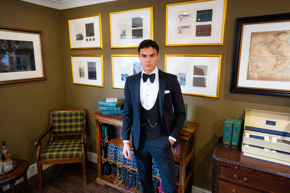 Slim fit classic black dinner suit with traditional black tuxedo jacket, classic black dinner suit trousers and black low cut horseshoe waistcoat. Features a slim fit 1 button velvet dinner jacket with wide black satin peak lapel. Worn with white studded dress shirt with French cuff shirt and large tom ford style bow tie and white pocket square.