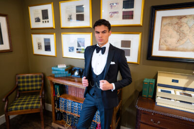 Slim fit classic black dinner suit in light weight, 100% wool. 3 piece dinner suit with traditional black tux jacket, classic black dinner suit trousers and black horseshoe waistcoat. Features a slim fit 1 button black dinner jacket with classic black satin peak lapel. Worn with white studded dress shirt with French cuff shirt and large tom ford style bow tie and white pocket square.