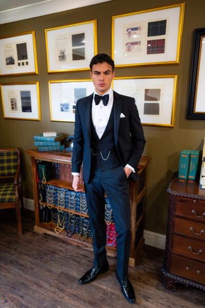 Slim fit classic black dinner suit with traditional black tuxedo jacket, classic black dinner suit trousers and black low cut v shape waistcoat. Features a slim fit 1 button velvet dinner jacket with wide black satin peak lapel. Worn with white studded dress shirt with French cuff shirt and large tom ford style bow tie and white pocket square.