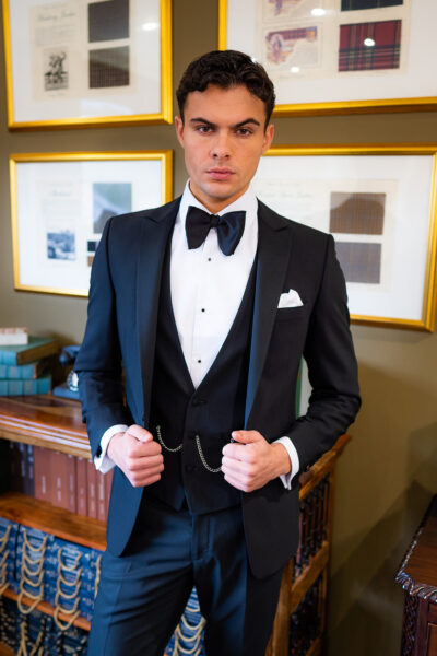 Slim fit classic black dinner suit with traditional black tuxedo jacket, classic black dinner suit trousers and black low cut v shape waistcoat. Features a slim fit 1 button velvet dinner jacket with wide black satin peak lapel. Worn with white studded dress shirt with French cuff shirt and large tom ford style bow tie and white pocket square.