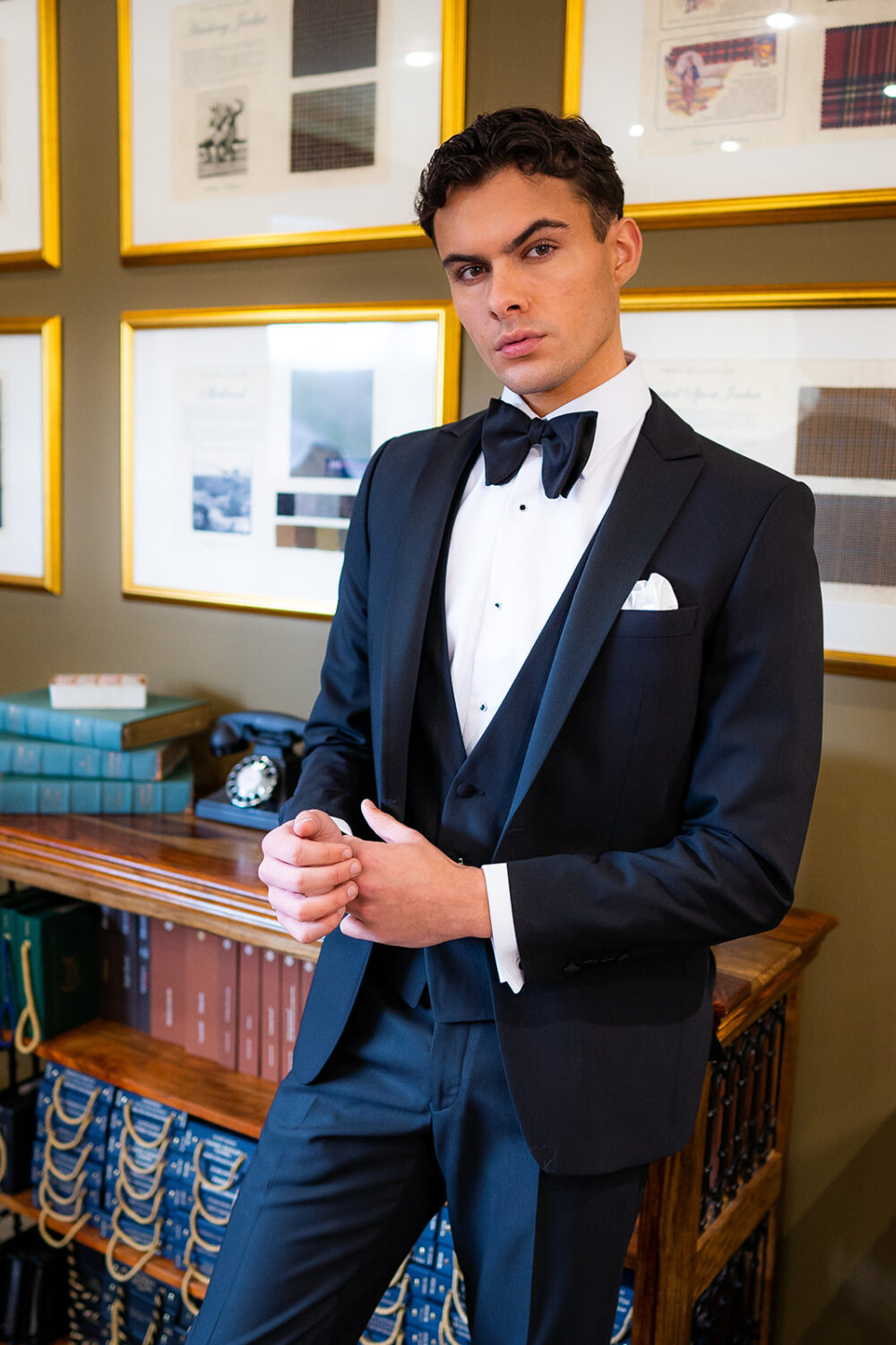 Slim fit classic black dinner suit with traditional black tuxedo jacket, classic black dinner suit trousers and black low cut v shape waistcoat. Features a slim fit 1 button velvet dinner jacket with wide black satin peak lapel. Worn with white studded dress shirt with French cuff shirt and large tom ford style bow tie and white pocket square.