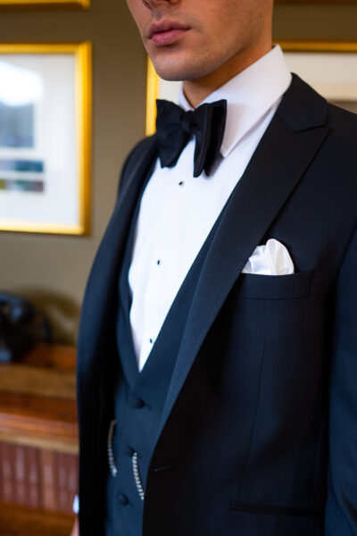 Slim fit classic black dinner suit with traditional black tuxedo jacket, classic black dinner suit trousers and black low cut v shape waistcoat. Features a slim fit 1 button velvet dinner jacket with wide black satin peak lapel. Worn with white studded dress shirt with French cuff shirt and large tom ford style bow tie and white pocket square.
