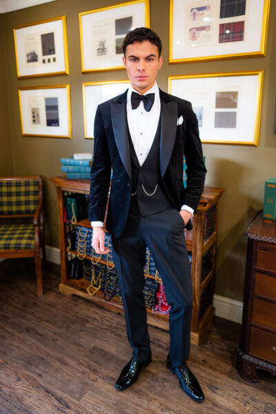 Slim fit classic black velvet dinner suit with traditional black velvet tux jacket, classic black dinner suit trousers and black low cut v shape waistcoat. Features a slim fit 1 button velvet dinner jacket with wide black satin peak lapel. Worn with white studded dress shirt with French cuff shirt and large tom ford style bow tie and white pocket square.