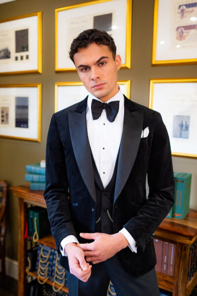 Slim fit classic black velvet dinner suit with traditional black velvet tux jacket, classic black dinner suit trousers and black low cut v shape waistcoat. Features a slim fit 1 button velvet dinner jacket with wide black satin peak lapel. Worn with white studded dress shirt with French cuff shirt and large tom ford style bow tie and white pocket square.
